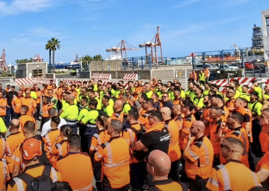 Trabajadores portuarios con ropa de alta visibilidad reunidos junto a instalaciones y grúas del puerto.